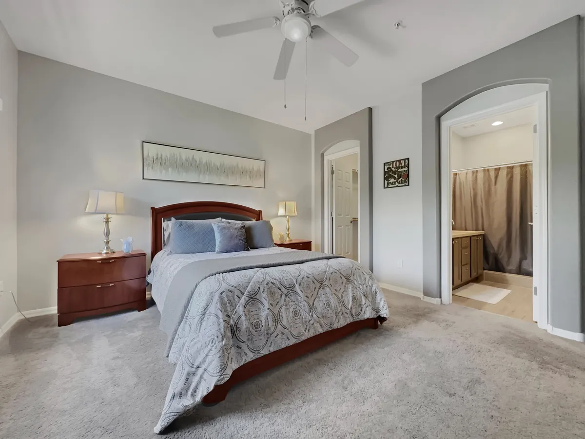 A serene bedroom with a plush queen-sized bed adorned in decorative bedding, a nightstand with a lamp for evening reading, tasteful artwork above the bed, and a doorway leading toward the bathroom. The room is softly lit, emphasizing the tranquil atmosphere and inviting relaxation.