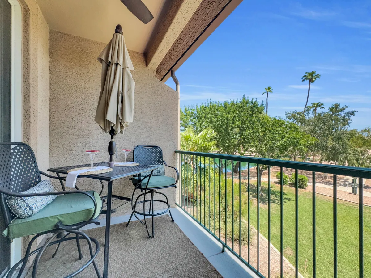 A cozy bistro table setup sits beneath an umbrella, surrounded by lush greenery. Drinks are set on the table, and the intimate, inviting arrangement highlights a casual, private outdoor experience perfect for morning coffee or sunset relaxation. The greenery in the background hints at a golf course or landscaped grounds.