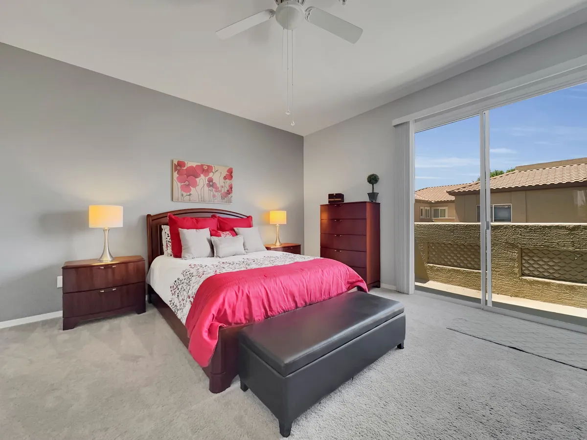 A bright bedroom featuring a queen-sized bed with red and white bedding, two bedside lamps for ambient lighting, framed artwork on the wall, and a large window that lets in natural morning light with views of the surrounding area.