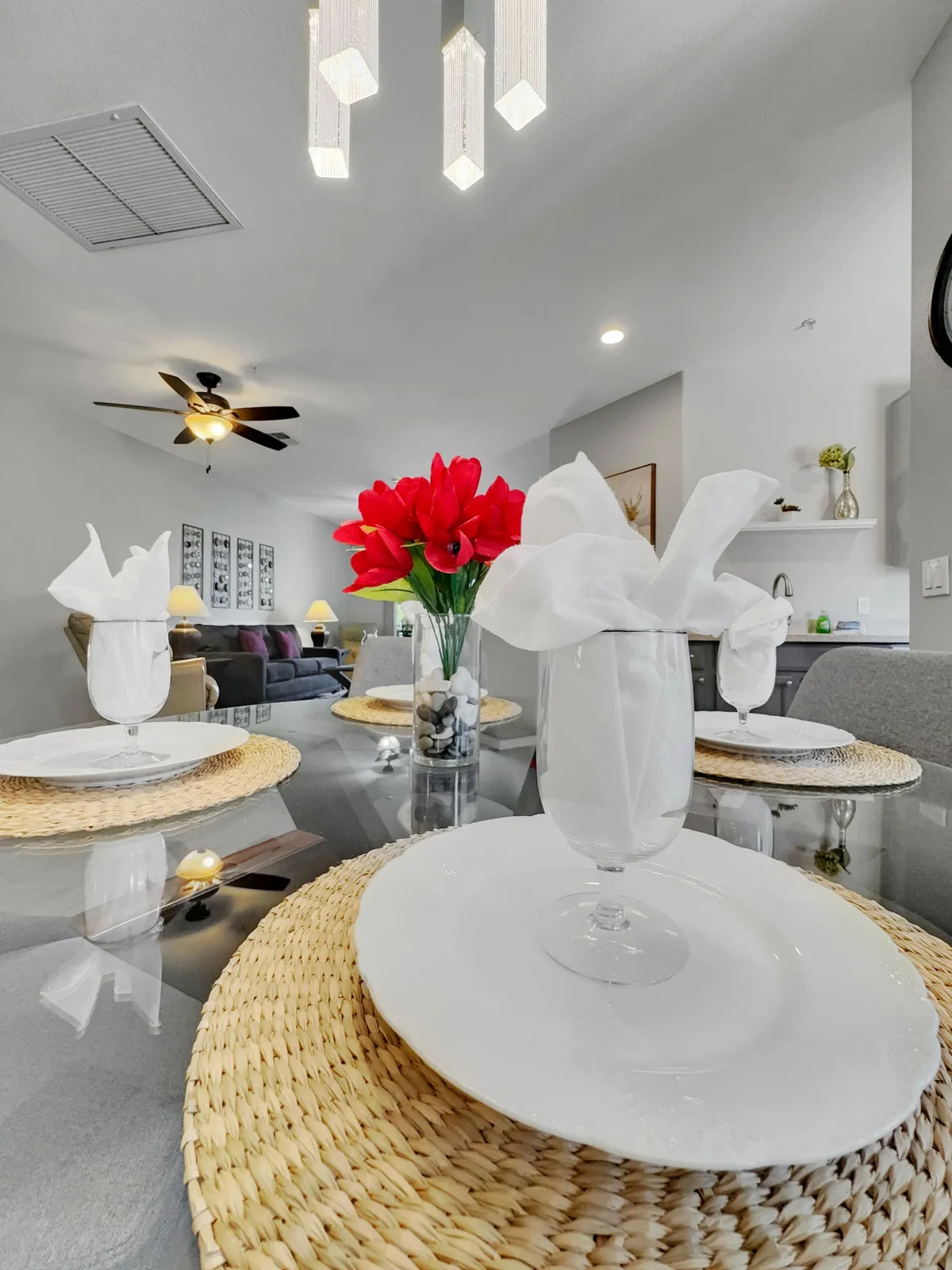 A warmly lit interior dining space with a stylish table setting, vibrant red flowers as the centerpiece, a chic chandelier overhead, and woven placemats, suggesting cozy meals and inviting gatherings.
