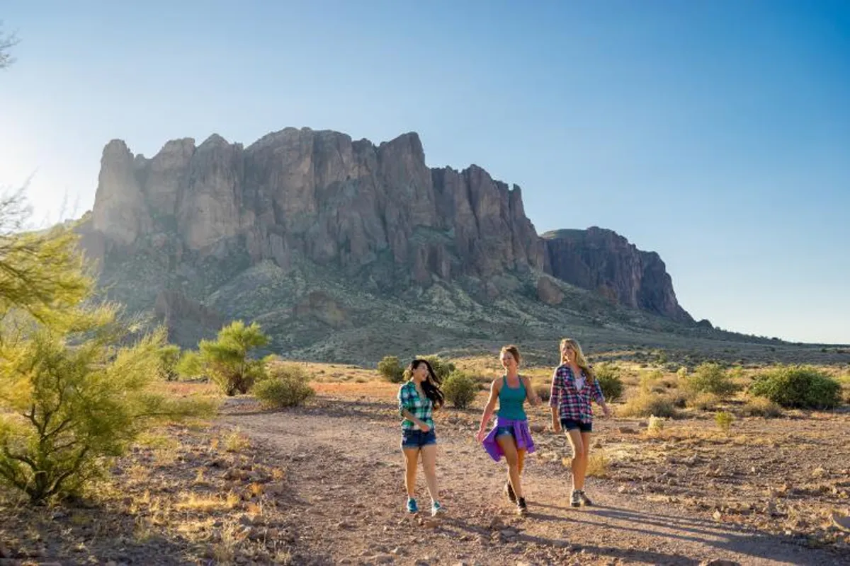 This exterior image features three women walking together through a scenic desert landscape, with rugged mountain backdrops and sun-drenched open spaces. The atmosphere is vibrant and joyful, emphasizing the connection to nature and the expansive beauty of Arizona.